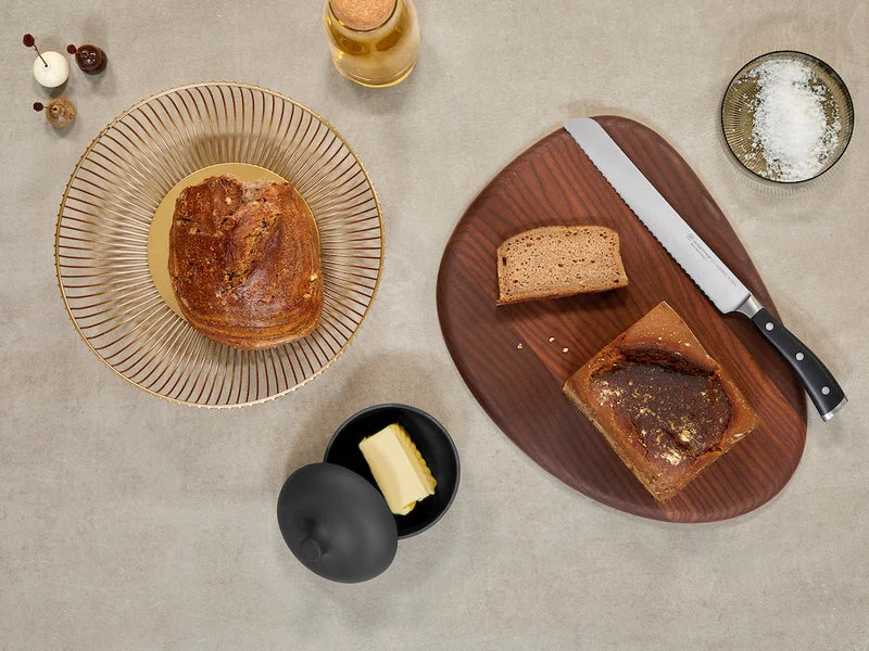 Serrated knife with black handle resting on a wooden cutting board beside sliced brown bread, a plate with a whole bread loaf, butter in a black dish with lid, a bowl of coarse salt, and a glass bottle on a beige countertop.